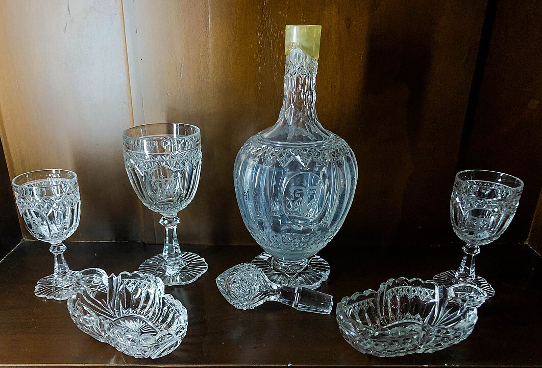 Crystal decanter set, ornate glass bottle with matching goblets, vintage cut glass drinkware, decorative glassware on wooden shelf, elegant display of antique crystal items