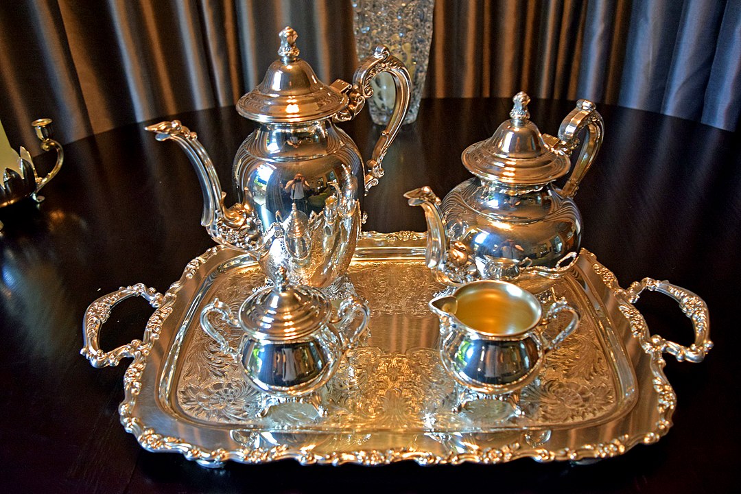 Silver tea set on ornate serving tray, includes teapot, coffee pot, sugar bowl, and creamer, placed on a dark wooden table, elegant and reflective surface, formal setting with curtain backdrop