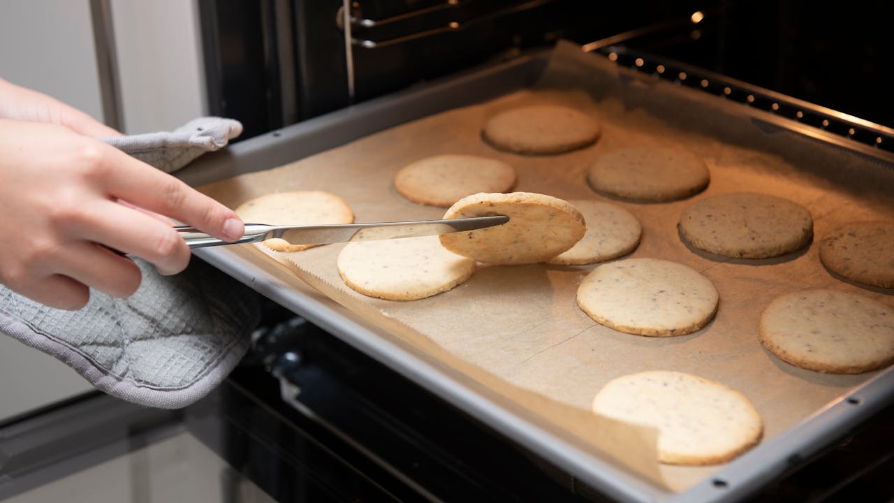 Person using tongs to lift a round cookie from parchment-lined baking sheet, cookies baking evenly in oven