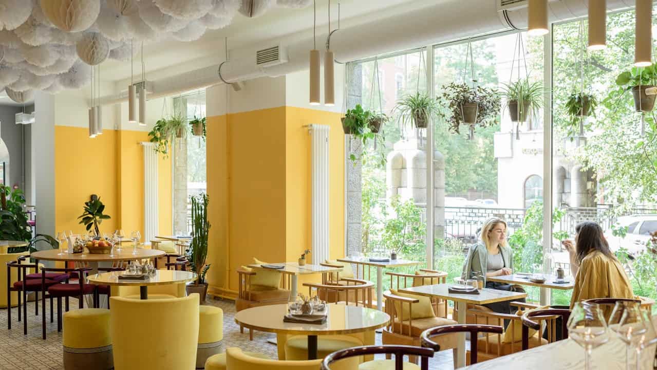  A brightly lit modern café interior with bold yellow walls and matching yellow chairs, round tables, hanging plants by the large windows, and decorative white paper clouds on the ceiling