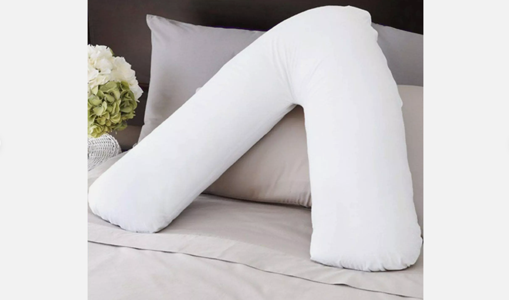 Boomerang-shaped white pillow placed upright on a neatly made bed with grey sheets and a floral arrangement beside it