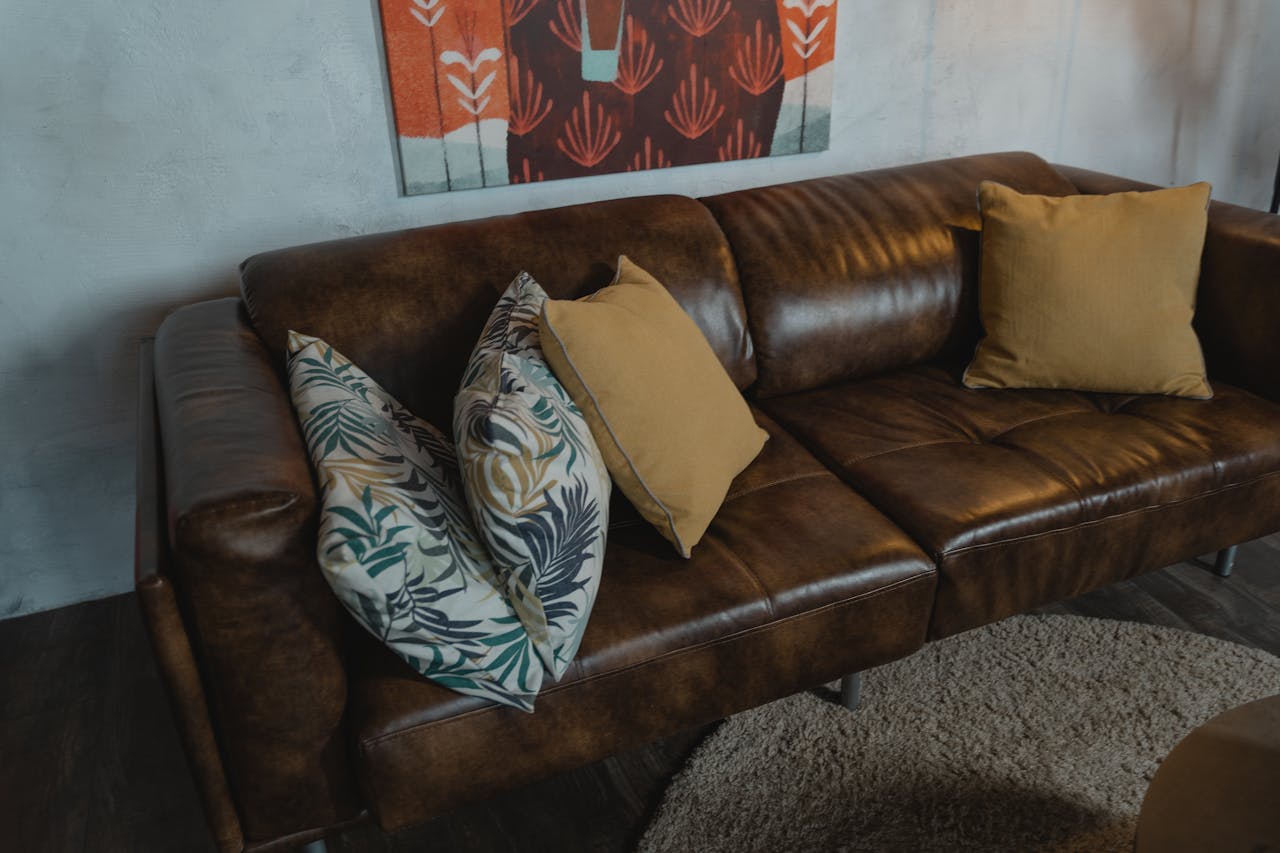 Brown brushed leather sofa with mustard and tropical leaf print throw pillows, set against a textured wall with modern red wall art