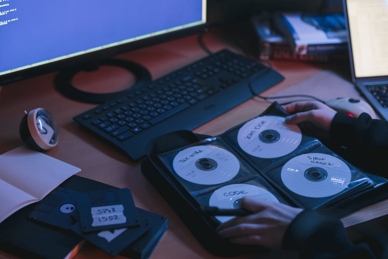 Person organizing labeled CDs in a black case, old floppy disks nearby, desktop computer setup in the background