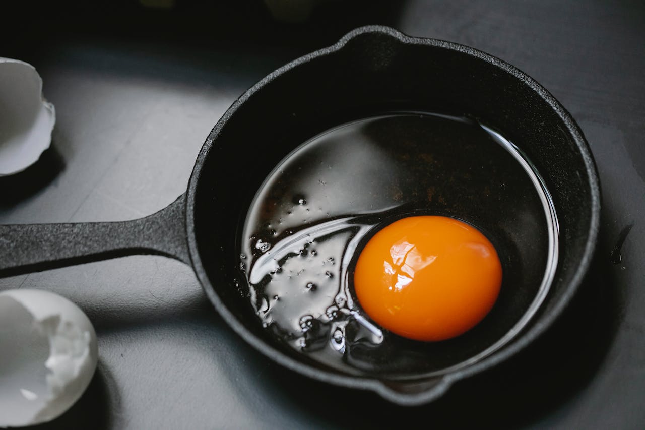 Raw egg with deep orange yolk in a black cast iron skillet, broken eggshells on nearby counter