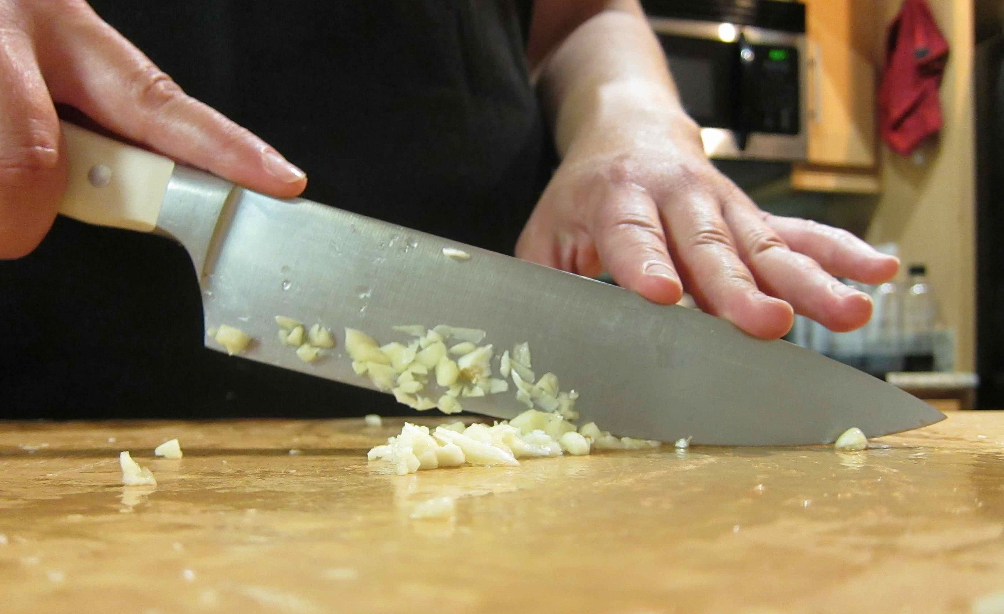 Close-up of a person mincing garlic with a stainless steel chef’s knife on a wooden cutting board in a kitchen