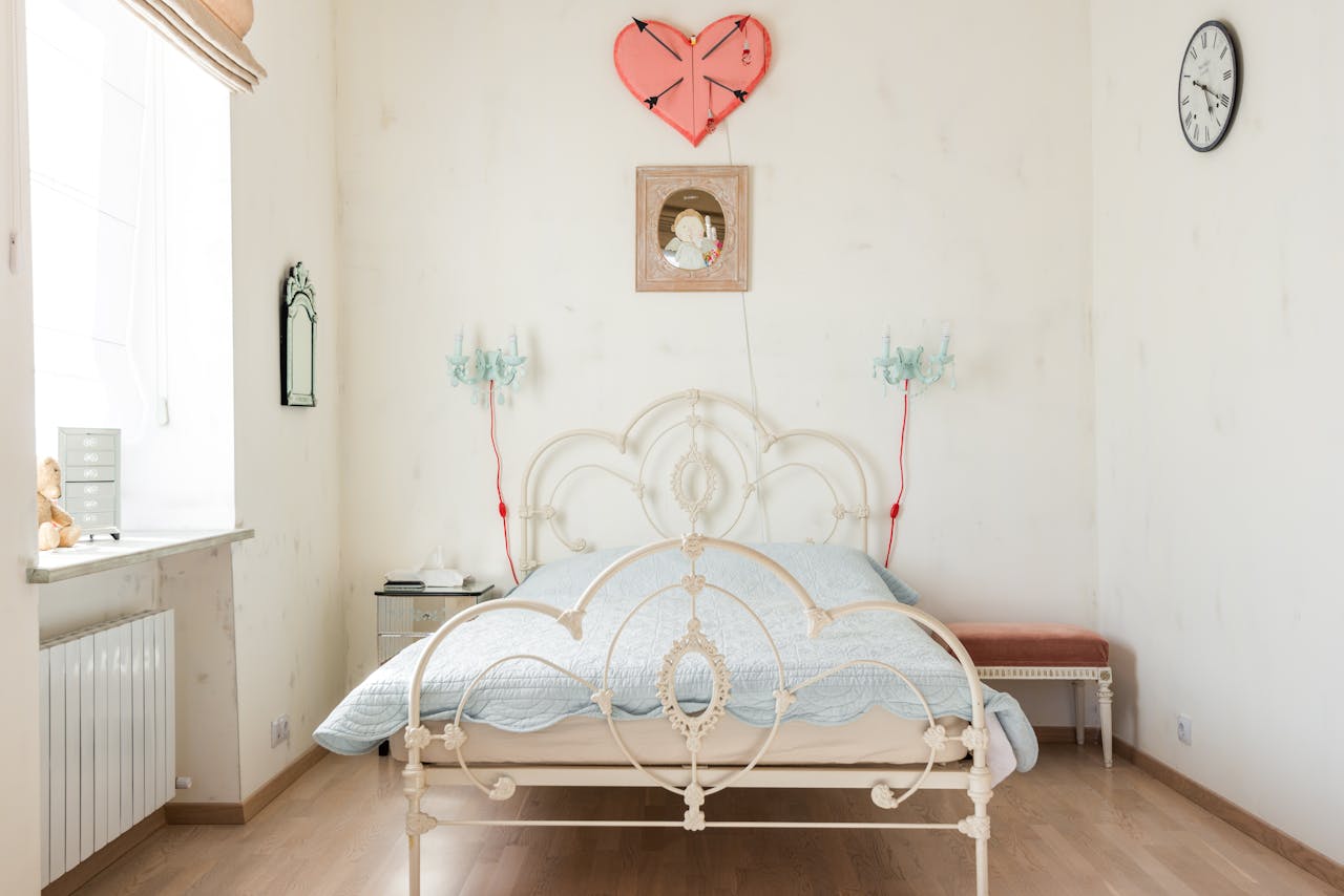 Vintage cream-colored metal platform bed with intricate scrollwork design, light blue bedding, and pastel wall decor in a bright bedroom