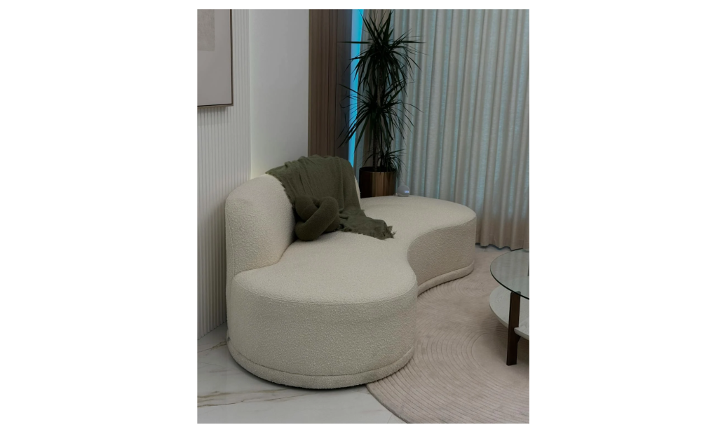 Modern living room with cream boucle curved sofa, green throw blanket and knot pillow, glass coffee table, neutral rug, potted plant, soft curtains