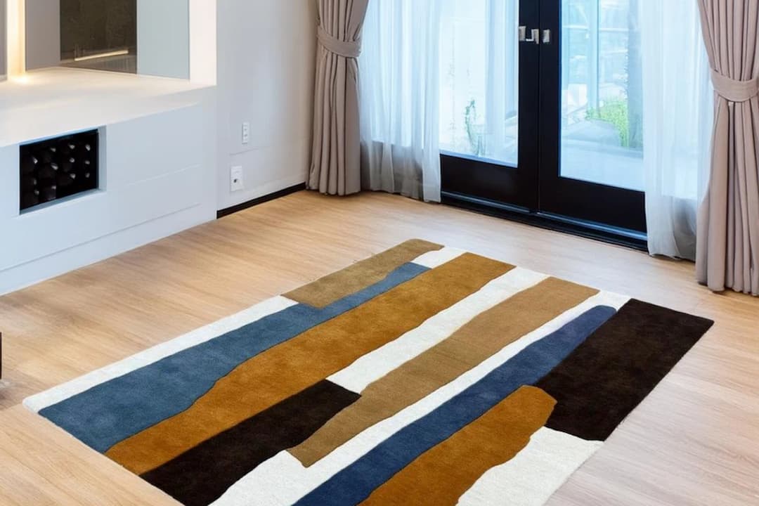  A modern living room with sheer curtains over large glass doors, featuring a rectangular area rug in earth tones including brown, navy, rust, and cream arranged in irregular vertical stripes
