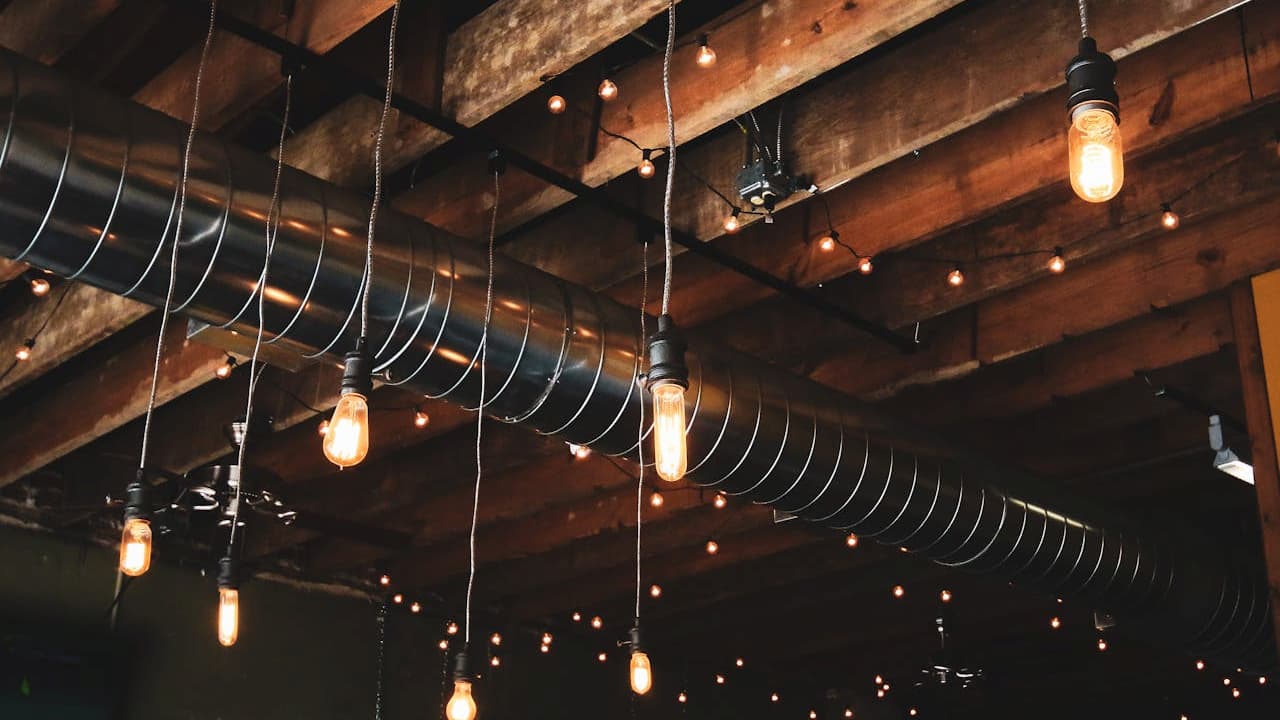 Industrial-style ceiling with exposed wooden beams, metal air ducts, and hanging Edison bulbs, enhanced by warm string lights creating a cozy ambiance