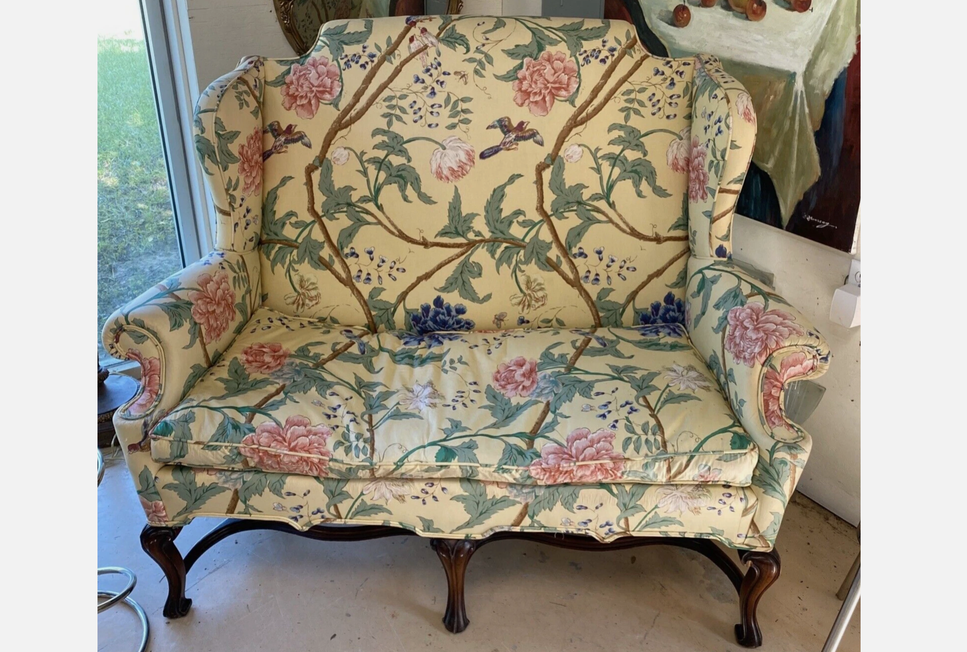 Vintage high-back sofa with cream-colored upholstery, floral and bird print, curved wooden legs, rolled arms, floral painting in background