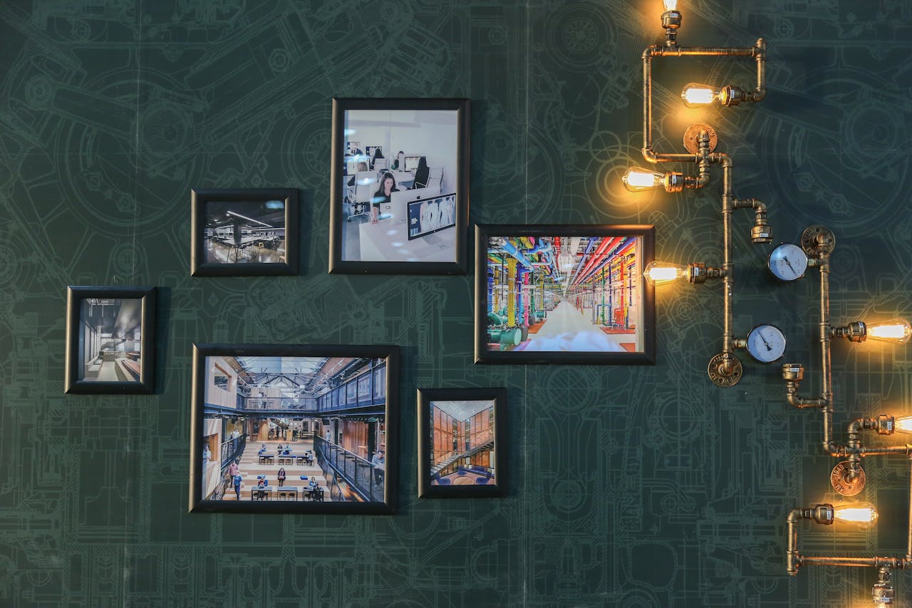 Gallery wall with framed industrial and architectural photos, dark green blueprint-patterned wallpaper, vintage-style pipe lighting with exposed bulbs and pressure gauges