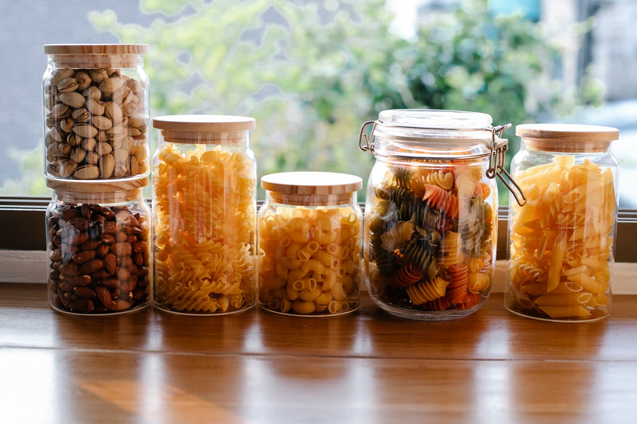 Glass jars filled with different dried pasta and nuts, placed on a wooden surface near a window