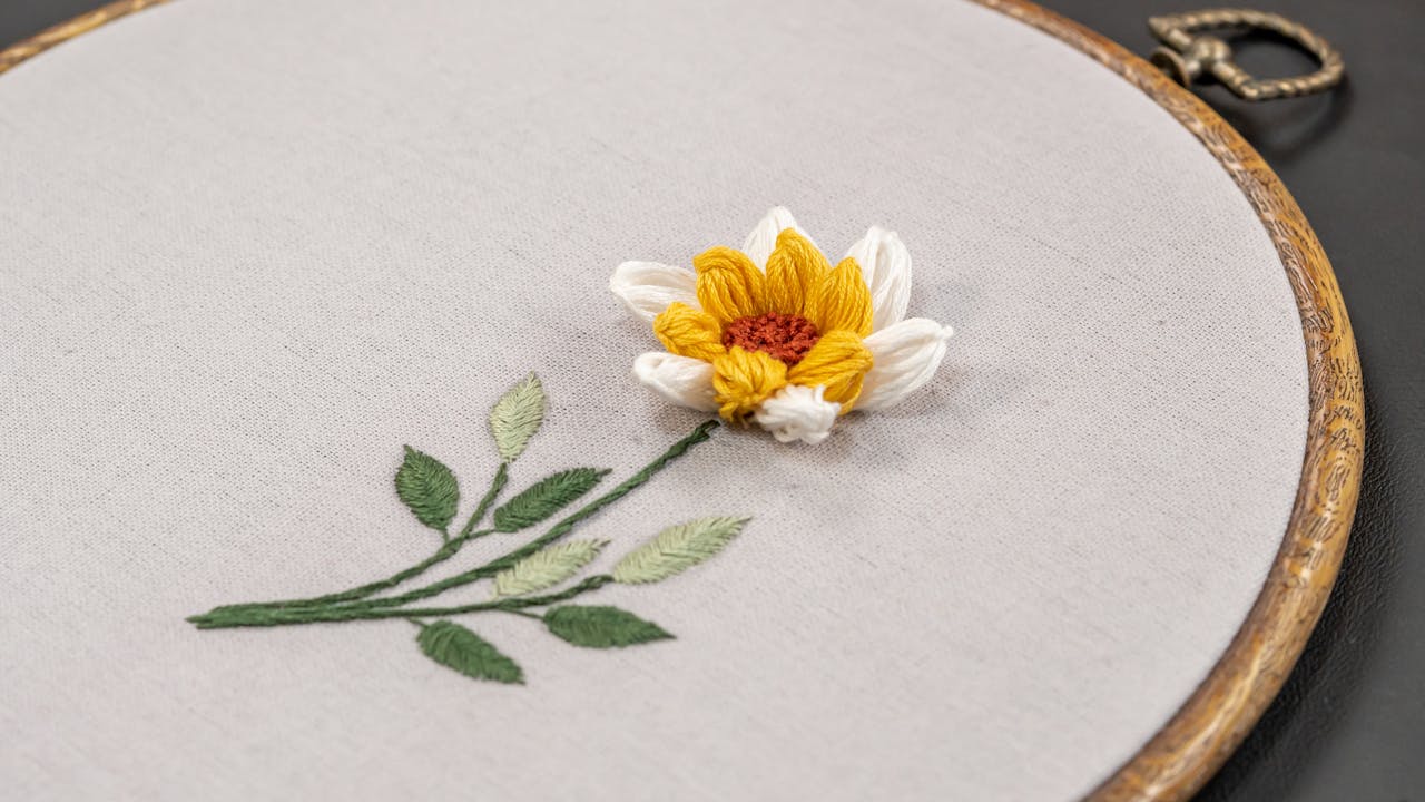 Hand-stitched embroidery of a single flower with yellow and white petals and green leaves on beige fabric in a wooden hoop