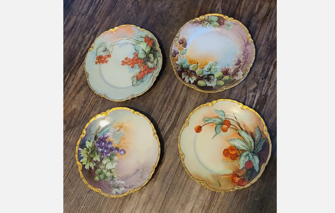 Four decorative porcelain plates, each with unique hand-painted floral and fruit designs, gold rims, placed on a wooden surface, arranged in a square pattern