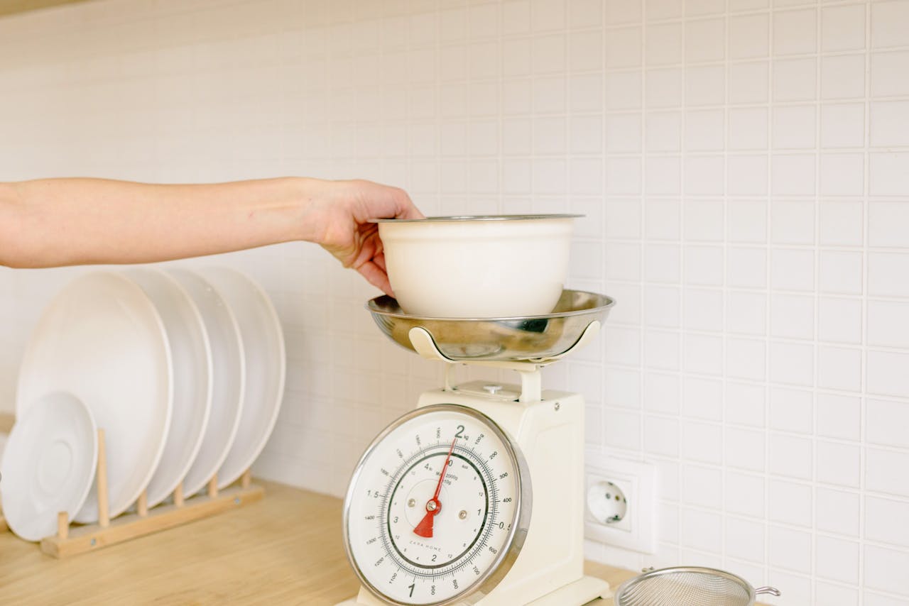 Hand placing a white bowl onto a retro-style analog kitchen scale, dishes drying in the background