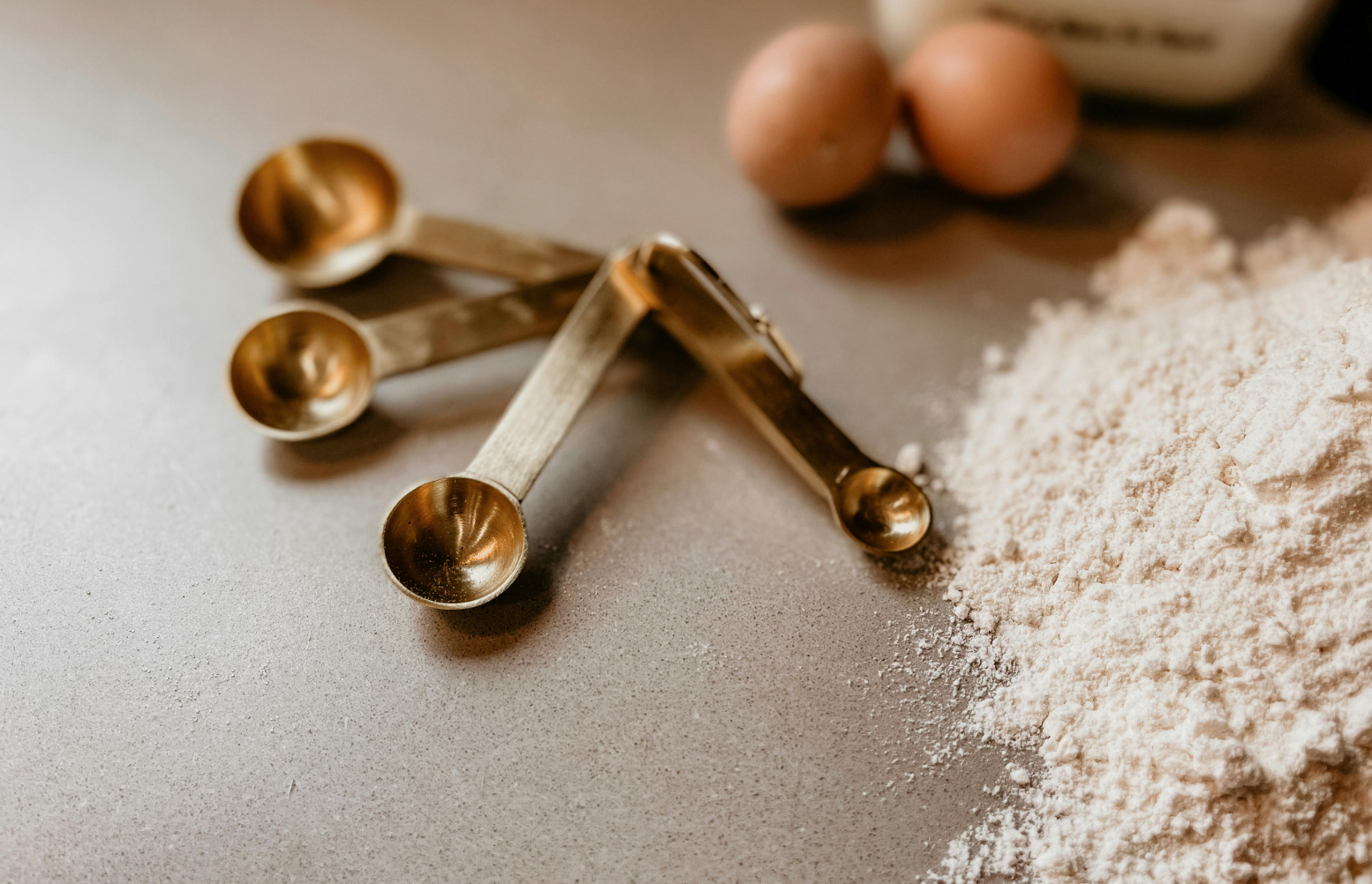 Set of brass measuring spoons on a kitchen counter beside a pile of flour and two brown eggs, with a baking ingredient container in the background