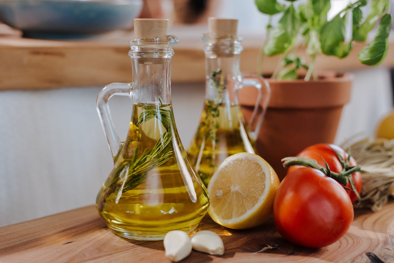 Two glass olive oil bottles with herbs and garlic, surrounded by lemon halves, tomatoes, and garlic cloves on a wooden surface