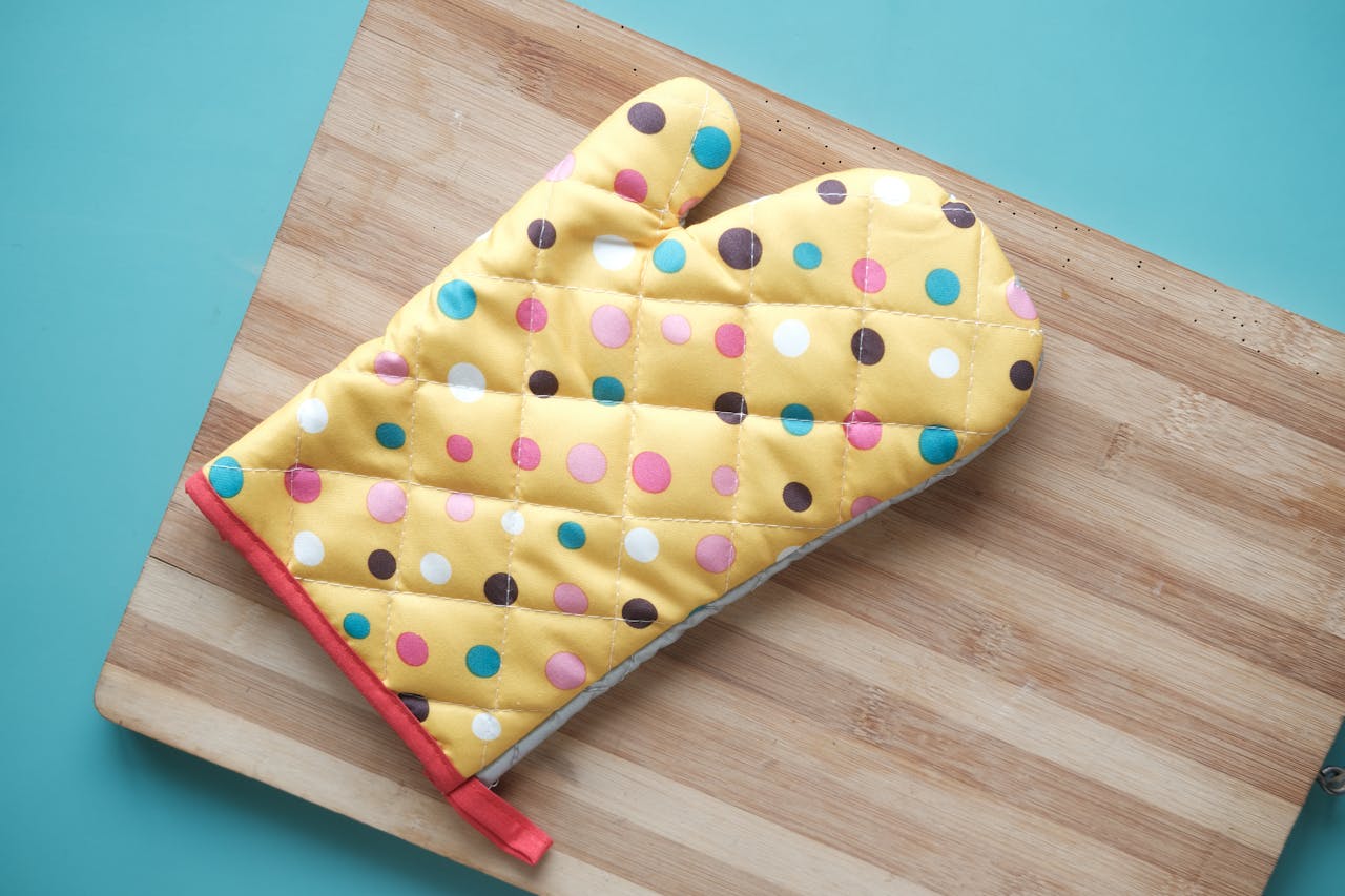 Yellow oven mitt with colorful polka dots resting on a wooden cutting board, background is a light blue surface