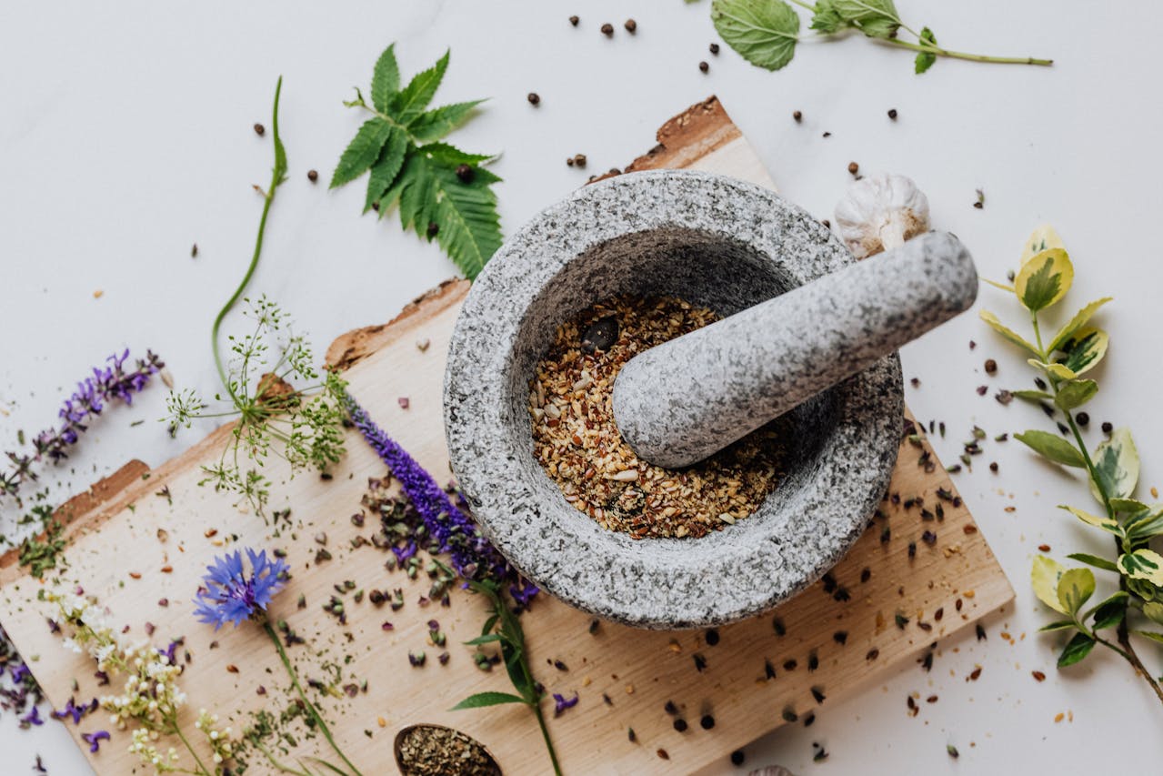 Granite mortar and pestle filled with crushed spices, surrounded by herbs and flower petals on a wooden board