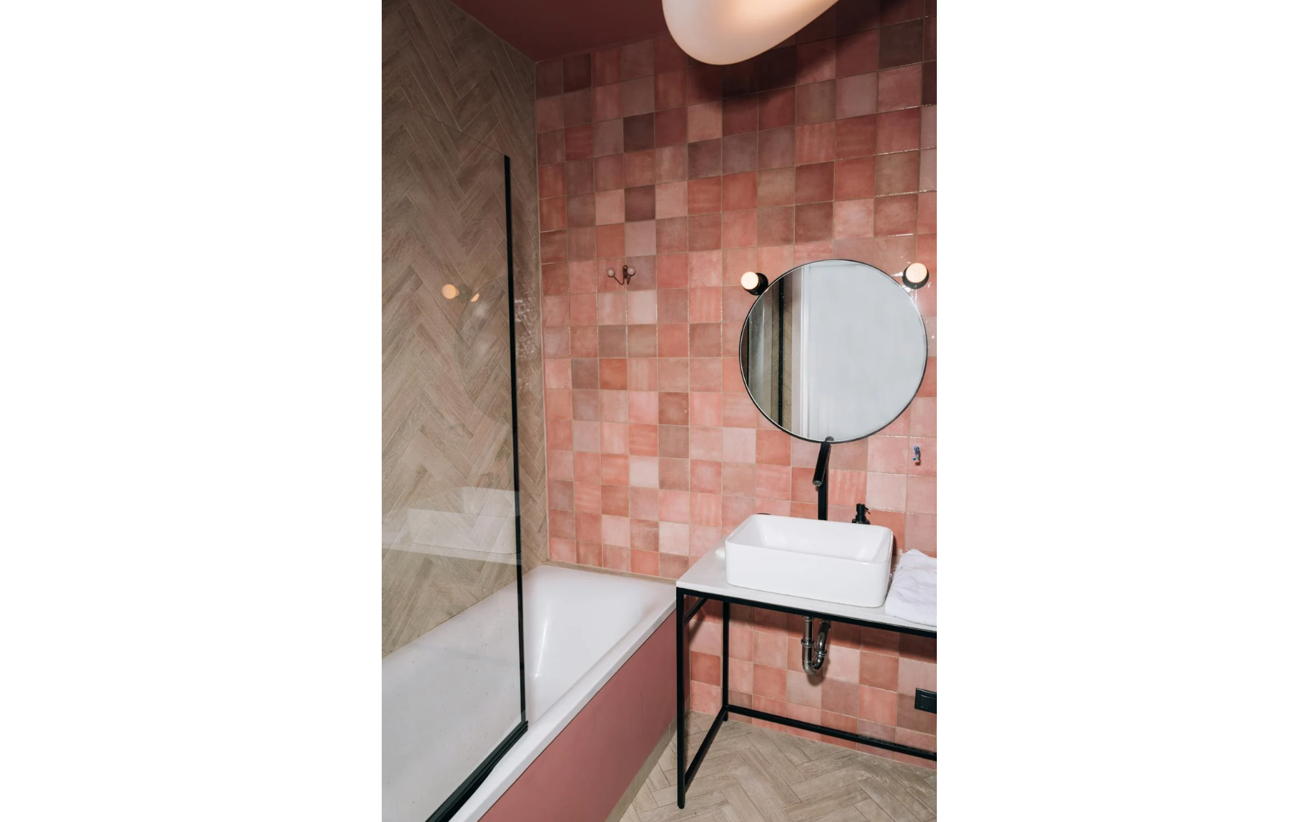 Bathroom with pink ceramic square tile wall, round mirror, wall-mounted black faucet, white vessel sink, bathtub with herringbone tile surround