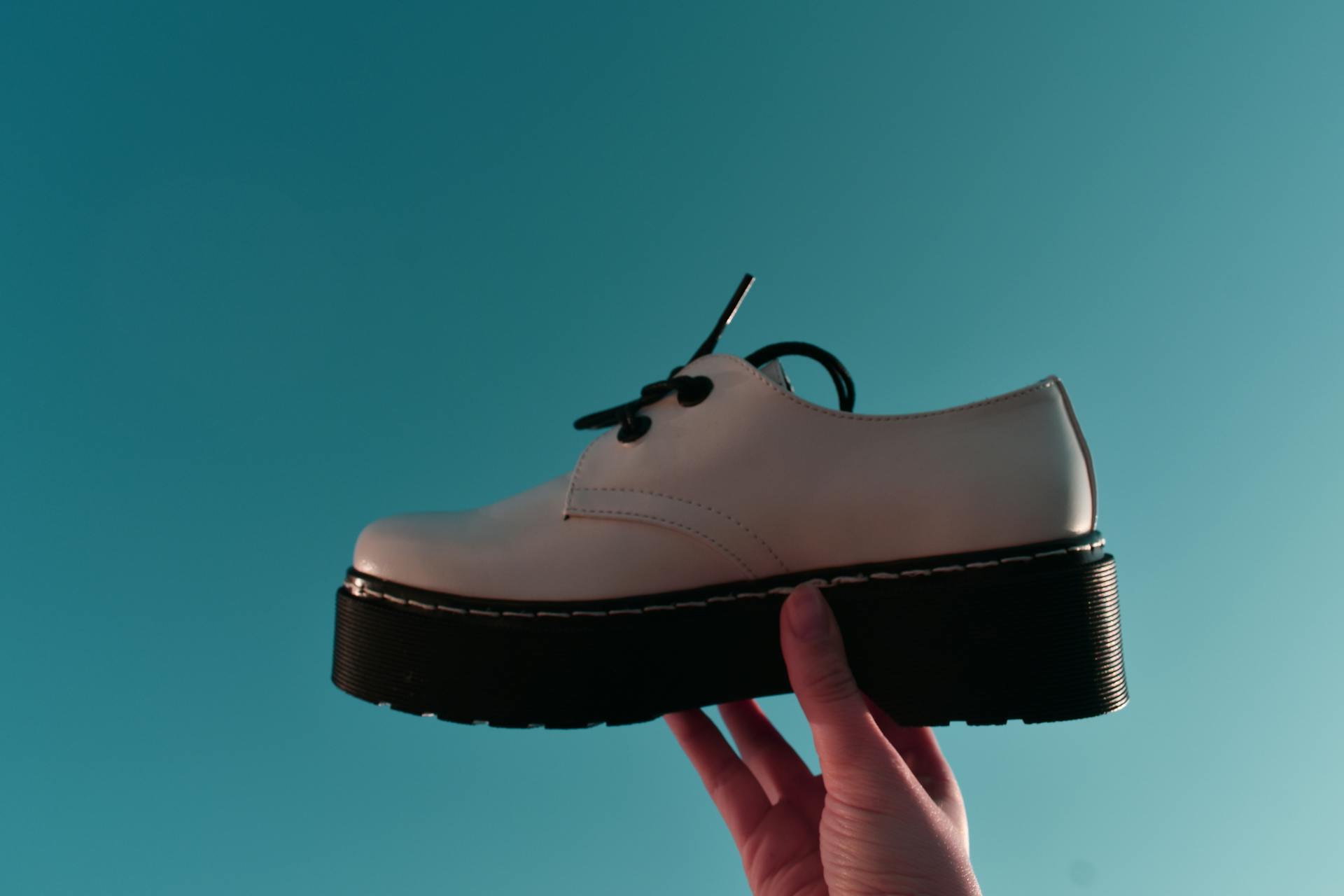 Platform shoes, White platform oxford shoe with black sole and laces held up by a hand against a teal blue background