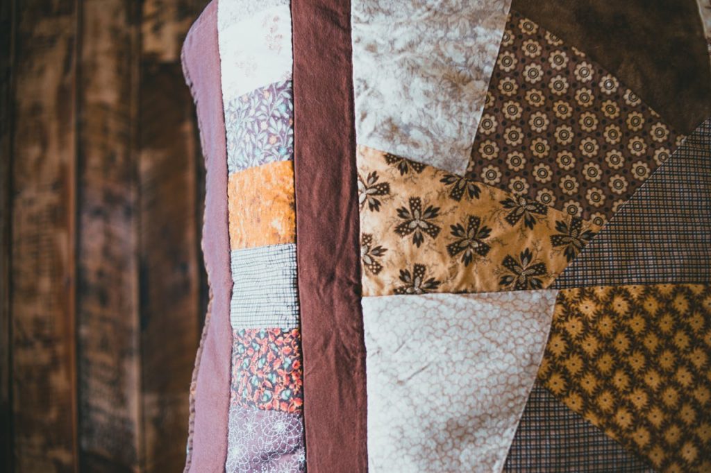 Close-up of a handmade patchwork quilt with floral and geometric prints in earthy tones, hanging in front of a rustic wooden wall
