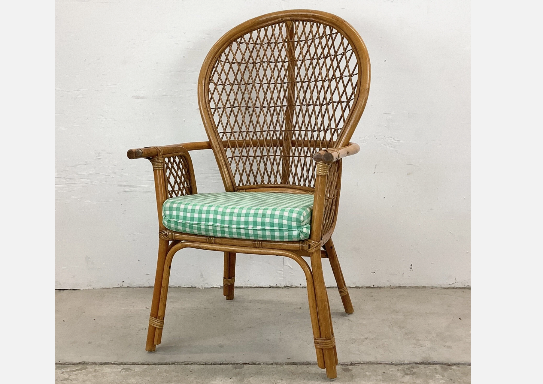 Vintage rattan armchair with curved high back and green gingham cushion, placed against a plain white wall
