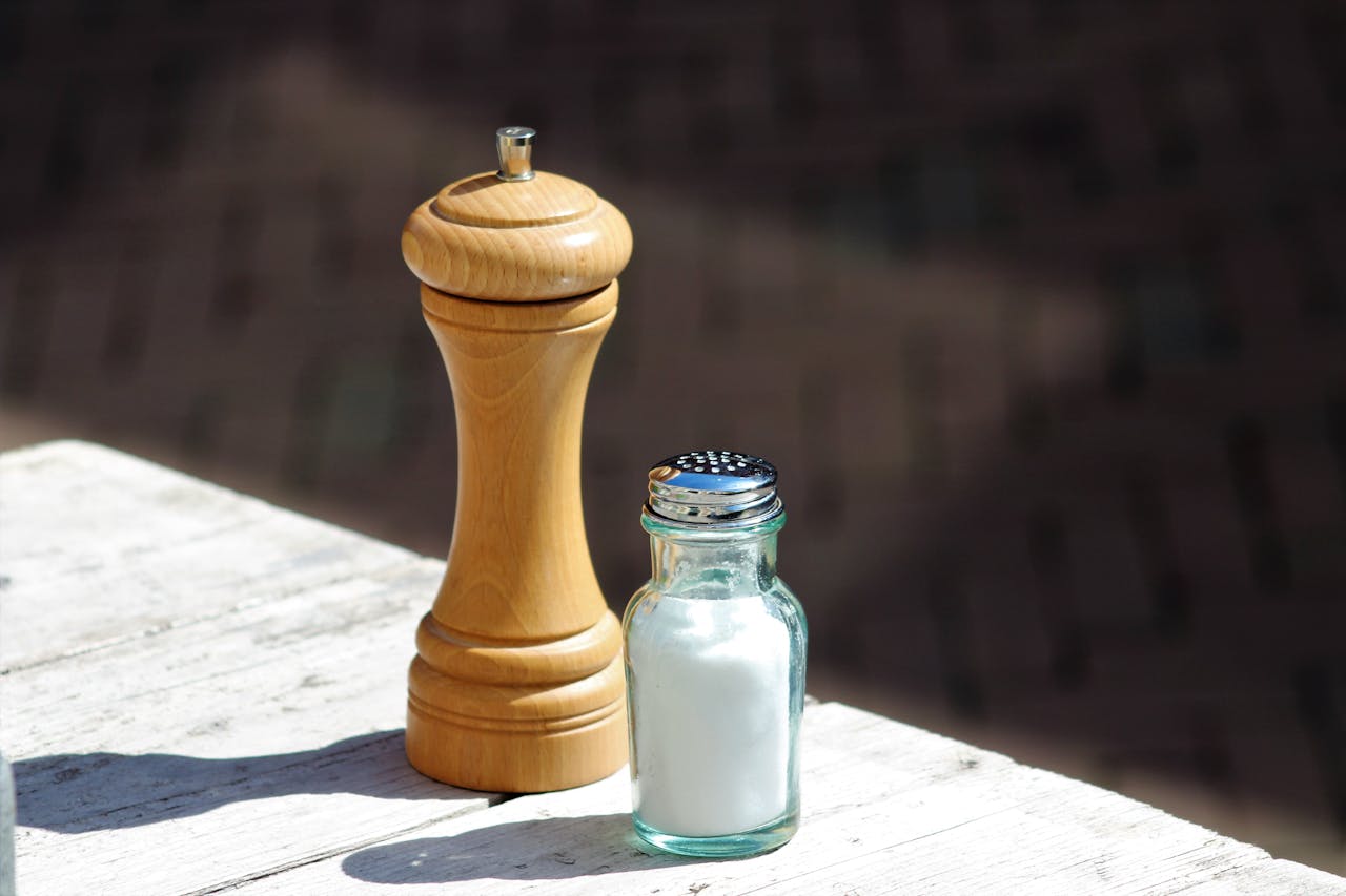 Wooden pepper grinder and glass salt shaker on a white wooden table in natural sunlight