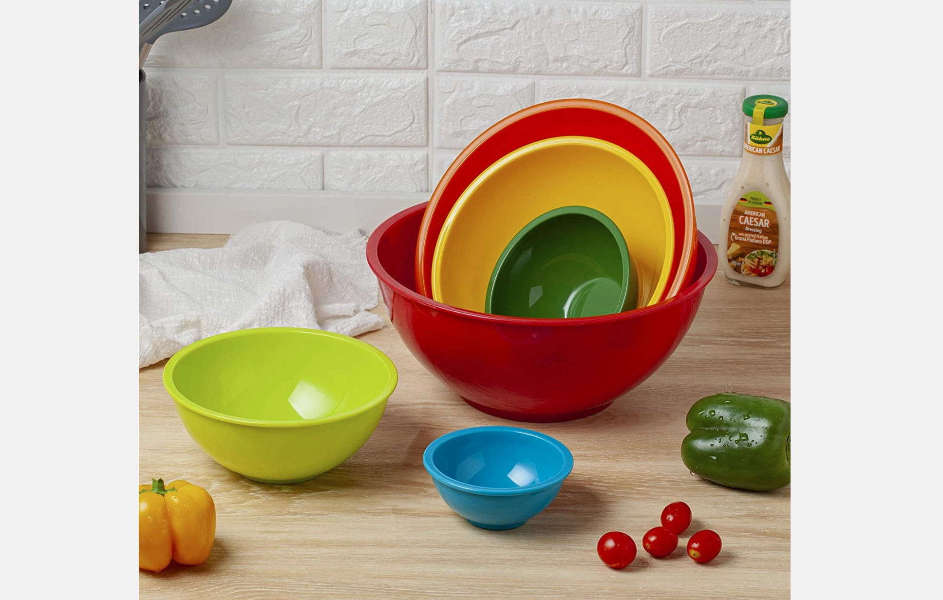 Set of colorful plastic mixing bowls in various sizes arranged on a kitchen counter, surrounded by fresh vegetables and a Caesar dressing bottle