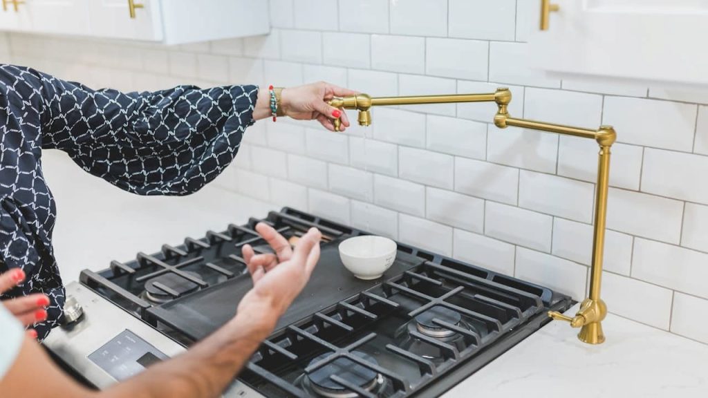 A person operates an unlacquered brass pot filler faucet above a stovetop with white subway tile backsplash and a small bowl beneath the water stream