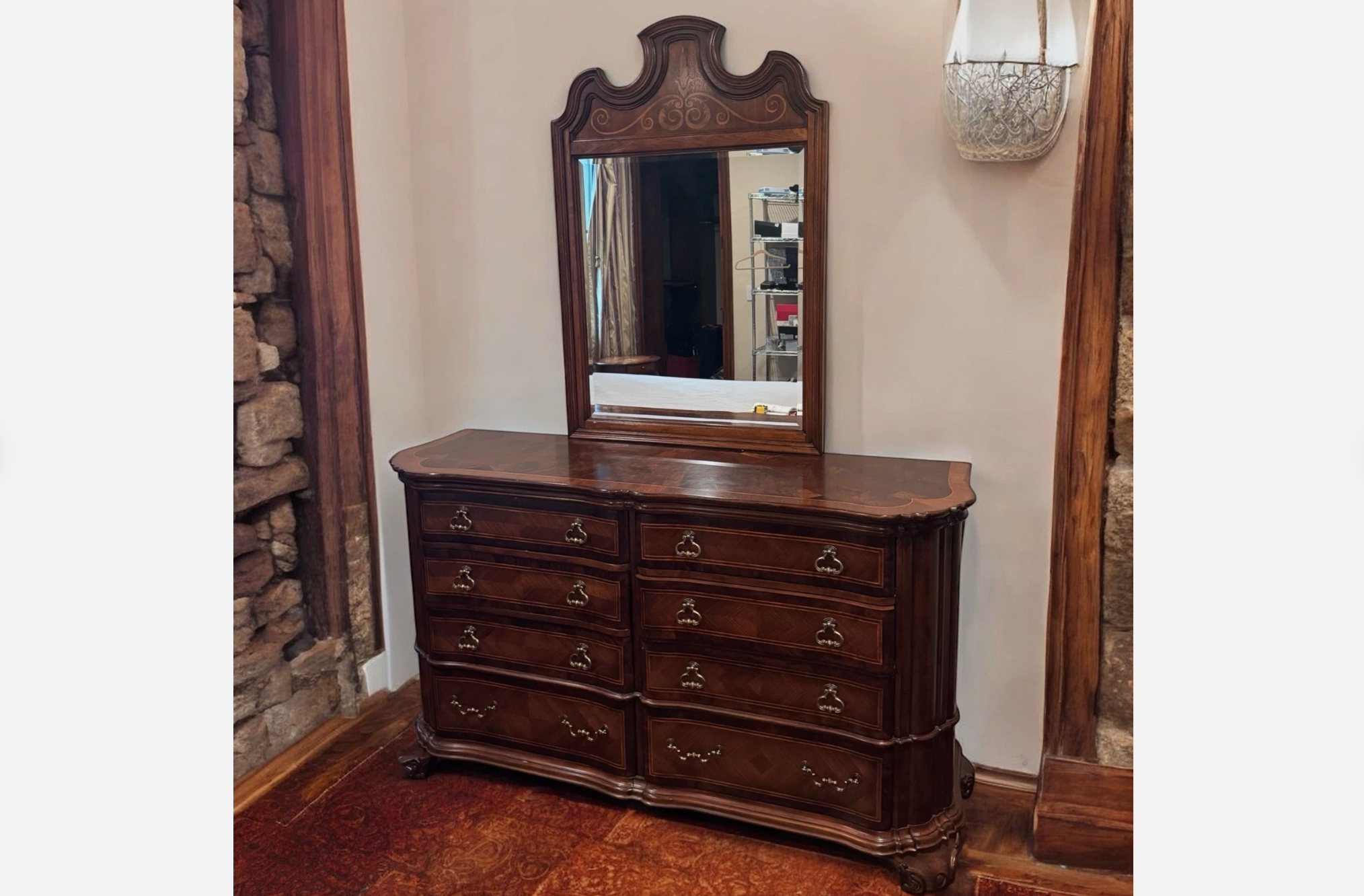 Ornate vintage dresser with curved drawers and matching carved mirror, rich wood finish, set against cream wall with rustic stone and wood frame accents