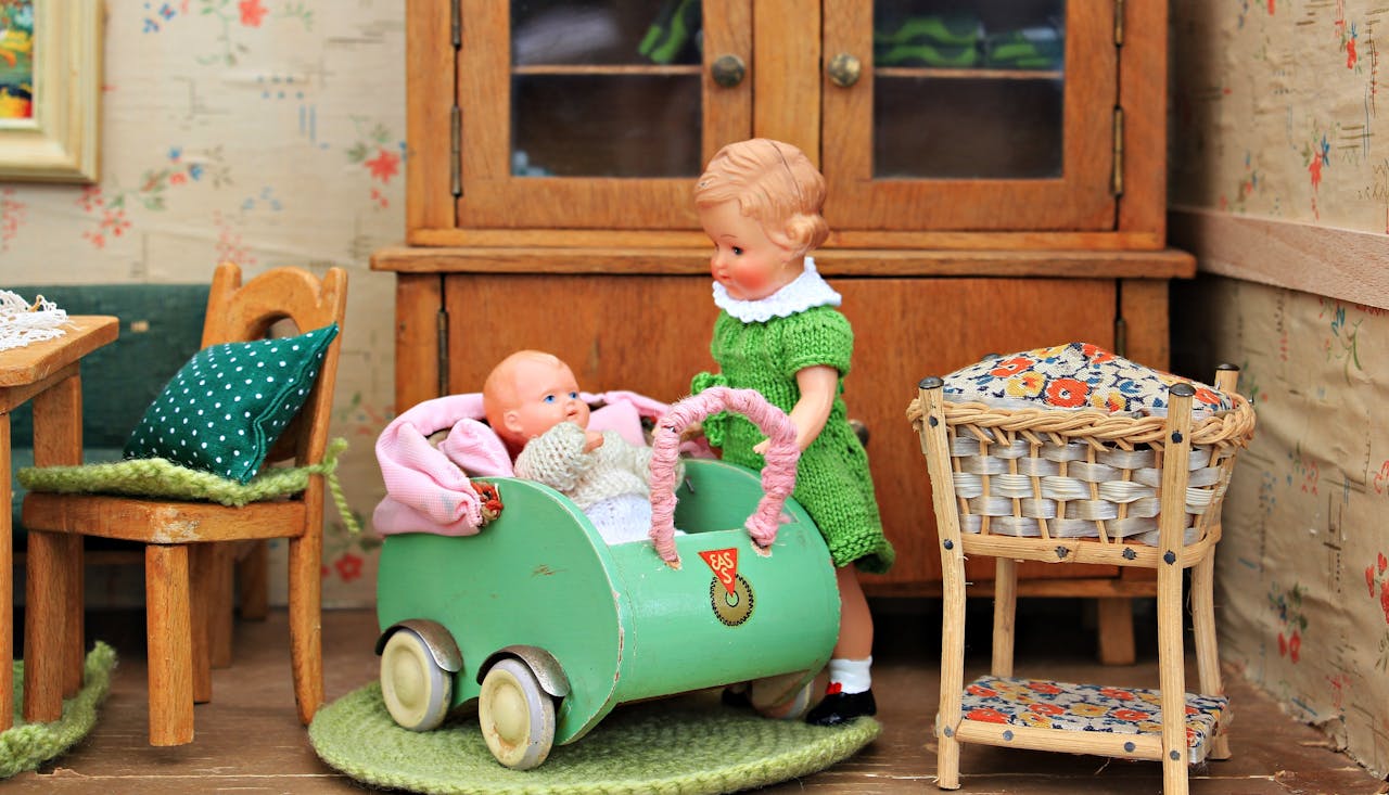 Vintage dolls in a dollhouse kitchen, one in a green dress pushing a baby doll in a pastel green stroller with wooden furniture