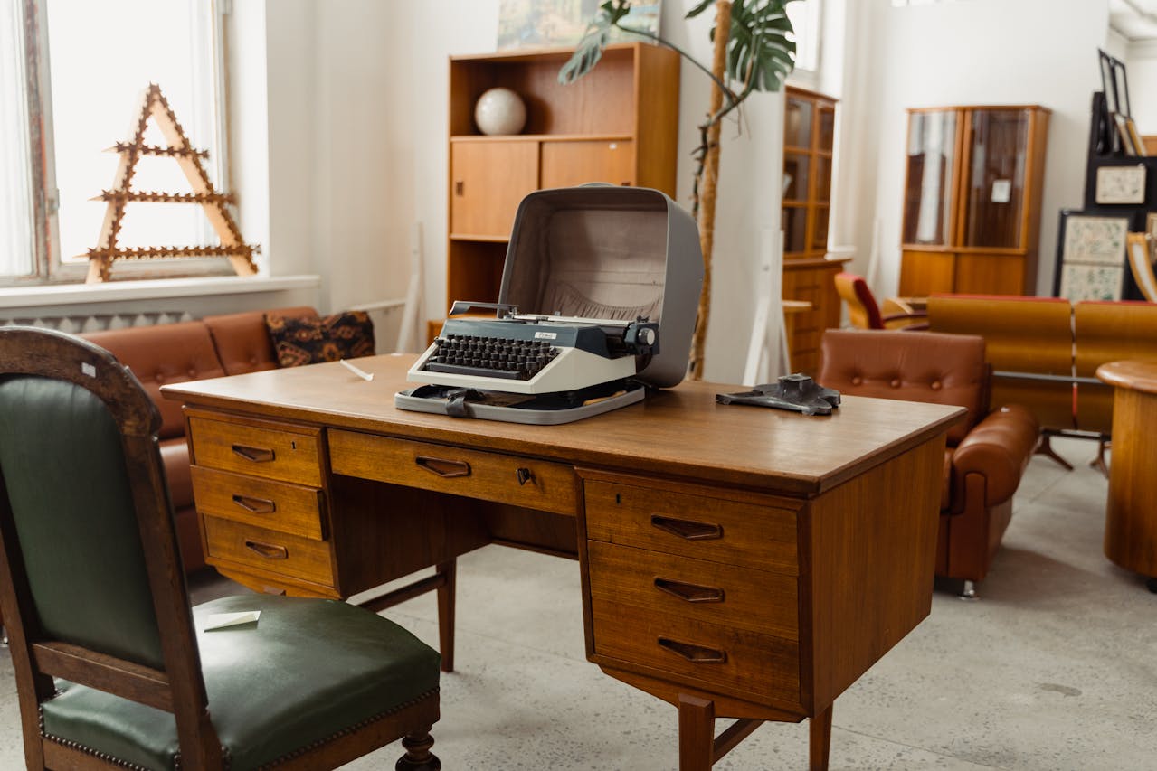 Vintage wooden writing desk with multiple drawers, an old typewriter in an open case on top, paired with a green leather chair