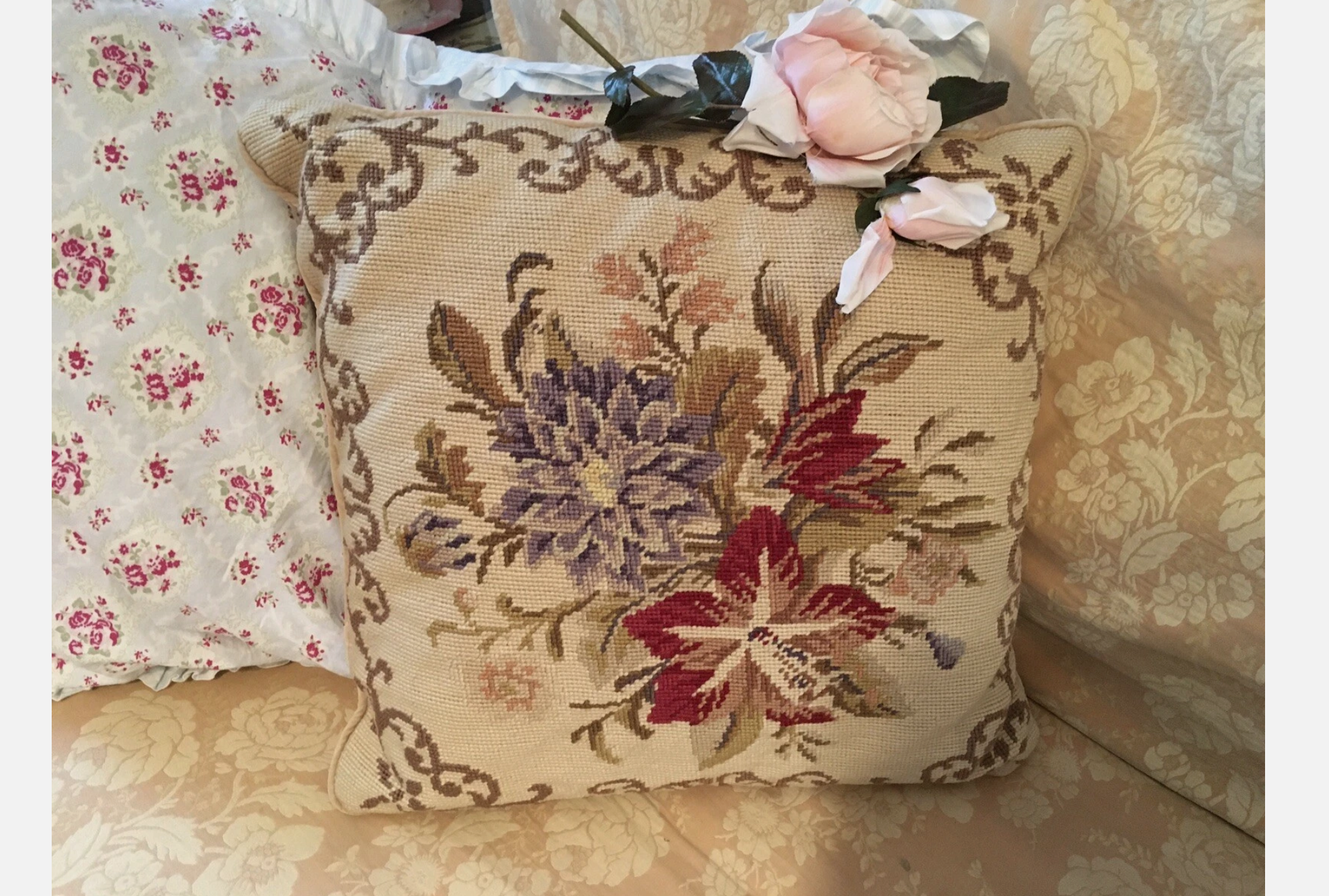 Vintage needlepoint pillow with floral embroidery in red, purple, and tan, resting on a floral-patterned sofa with a pink faux rose on top