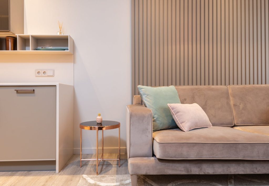 A modern living room with wood-paneled walls, a taupe velvet sofa, pastel throw pillows, and a small round copper-legged side table