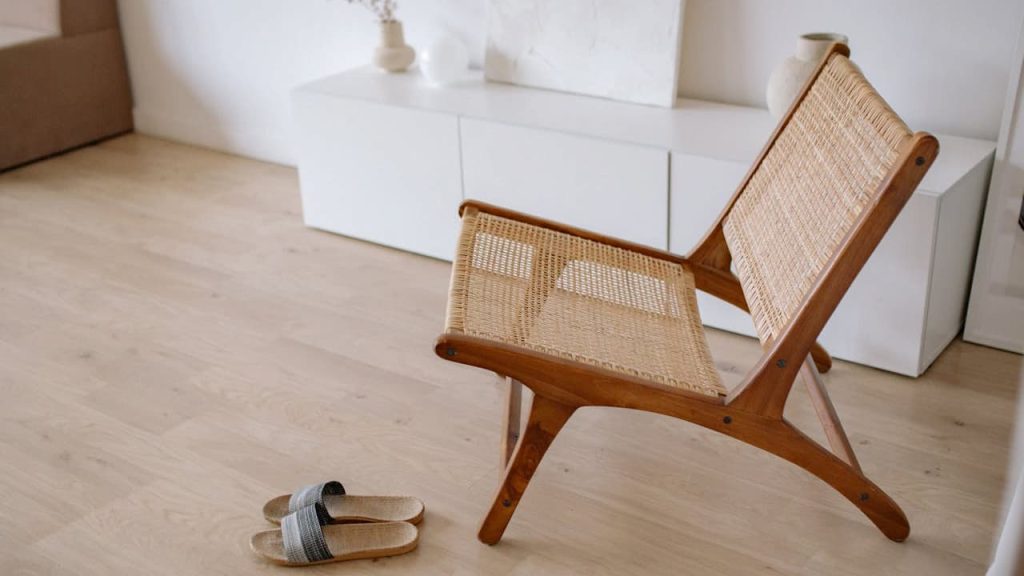 A woven rattan lounge chair with a wooden frame placed on a light wood floor in a minimalist living room, with a pair of woven slippers beside it and white cabinets in the background