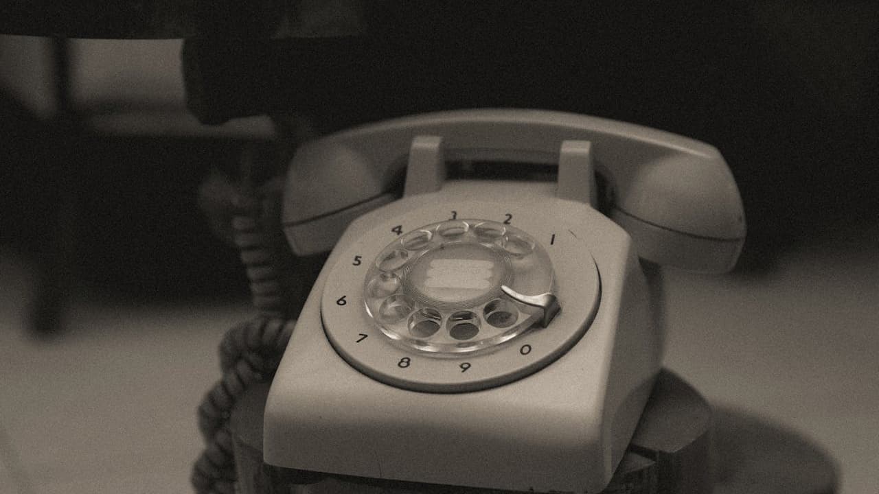 Vintage rotary dial telephone, light-colored body, coiled cord, placed on a table, with a dark, blurred background