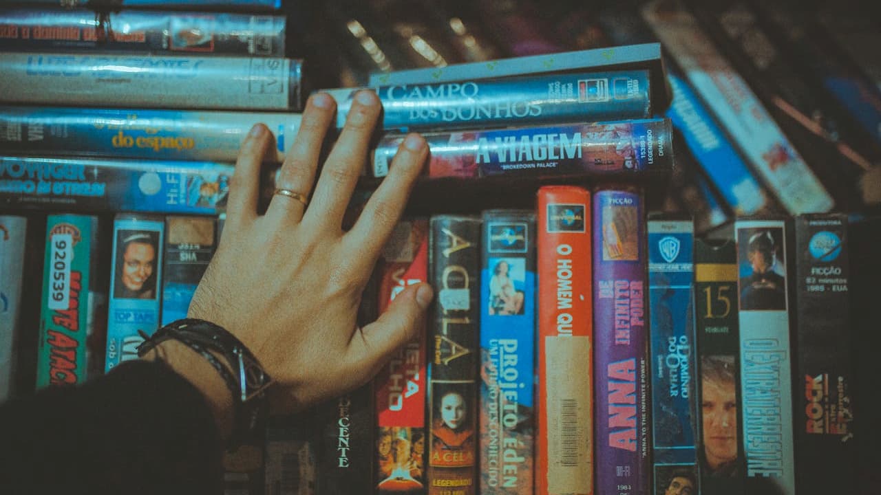 Hand reaching over a collection of colorful VHS tapes, wearing a black watch and silver ring, tapes stacked vertically and horizontally, with visible film titles and worn covers, suggesting a nostalgic or vintage movie collection