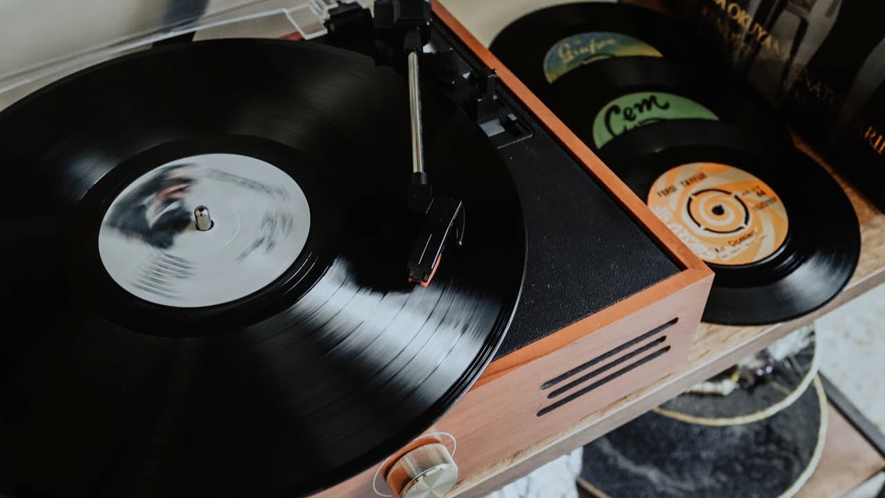 Close-up of a wooden turntable playing a black vinyl record, tonearm positioned on the spinning disc, additional records with colorful labels stacked nearby, vintage setup on a wooden shelf, cozy and nostalgic ambiance