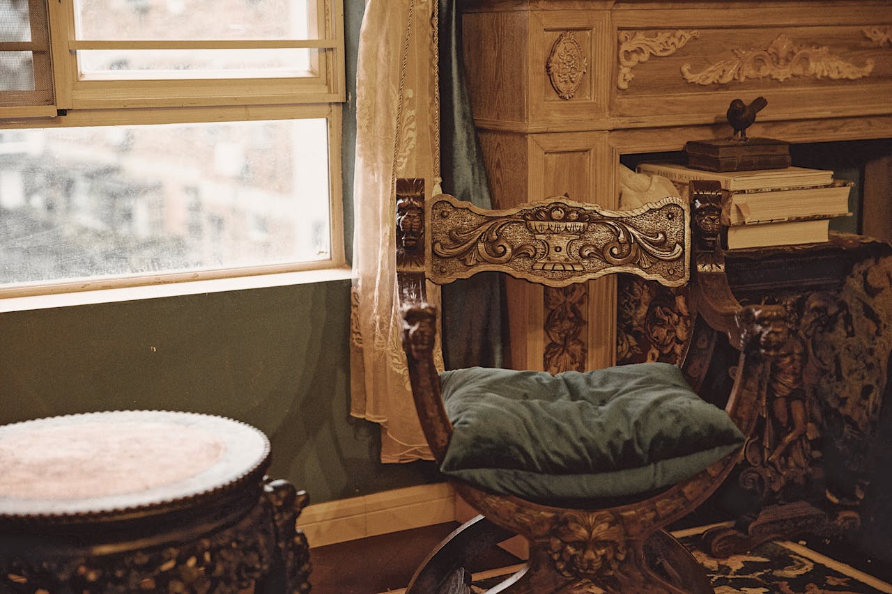 Intricately carved vintage wooden chair with green cushion, placed near window with sheer curtain, ornate furniture and stacked books in background