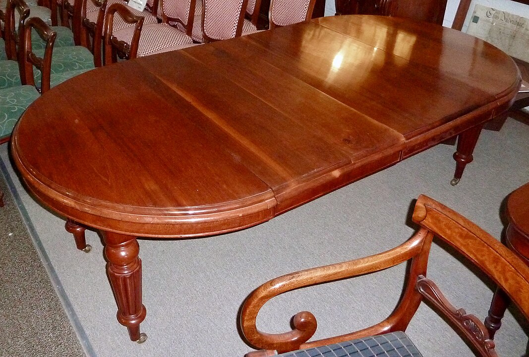 Large Victorian-style mahogany dining table with an oval shape and central leaf extension, supported by carved turned legs, surrounded by matching wooden chairs with curved armrests, placed on a carpeted floor.