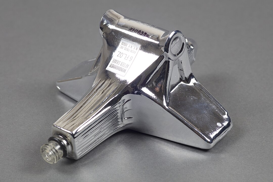 Chrome Avon bottle shaped like a jet or car, metallic finish, compact size, vintage collectible, novelty design, angled on a plain gray surface, retro aesthetic, reflective surface detailing