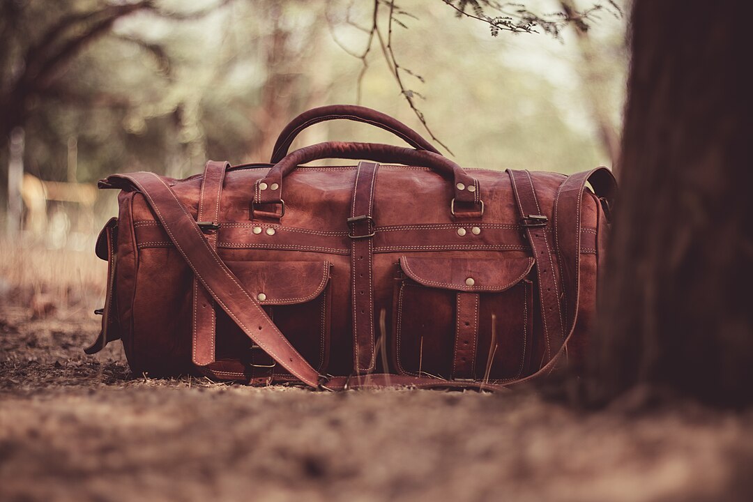 Brown leather duffel bag with multiple pockets, lying on the forest floor, surrounded by dry leaves and tree trunks, natural outdoor lighting, soft focus background with trees