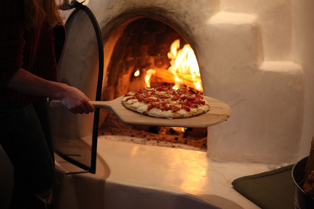 Person placing homemade pizza into wood-fired oven, pizza on wooden peel, visible fire burning inside rustic clay oven, cozy kitchen atmosphere, warm lighting, hand holding peel from left side, fresh toppings on pizza, curved oven opening, traditional cooking method