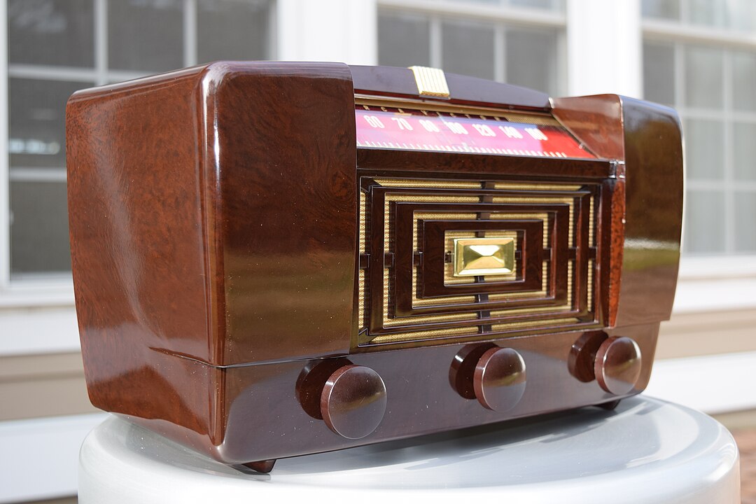 Vintage brown tabletop radio, glossy wooden casing, three control knobs at the bottom, rectangular speaker grille with gold detailing, displayed on a white surface, set against a window background
