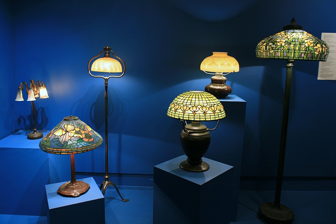 A display of vintage Tiffany-style lamps with stained glass shades, arranged on blue pedestals and against a blue wall, featuring various sizes and designs, including table and floor lamps, all warmly lit