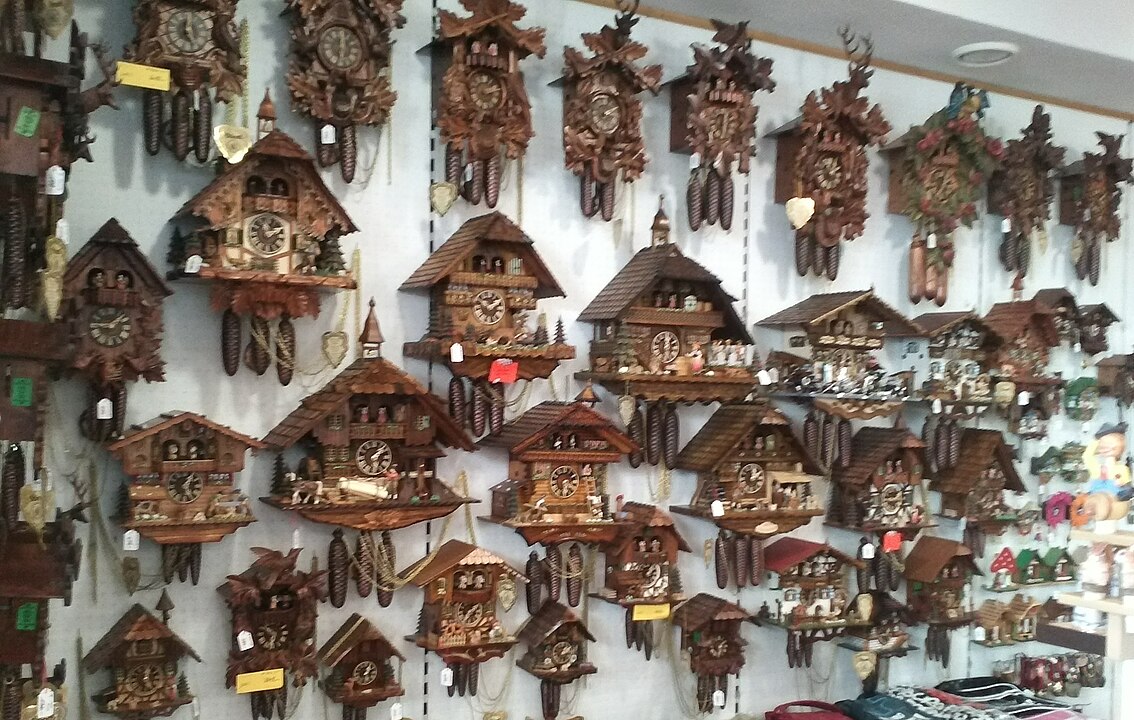 Wall display of traditional wooden cuckoo clocks, various sizes and designs, carved wooden details, hanging pendulums and weights, mounted on a white wall, arranged in rows, indoor shop or showroom setting