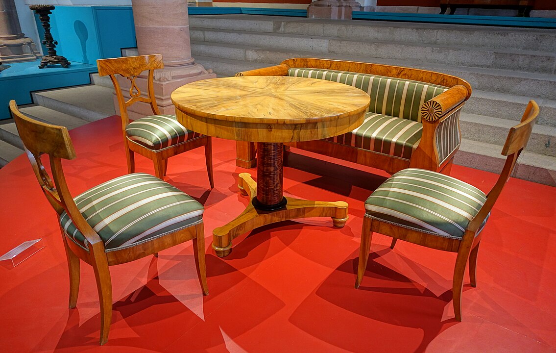 Biedermeier furniture set including a round wooden pedestal table, a barrel-back upholstered settee, and three matching wooden chairs with green and white striped fabric, displayed on a red platform in a museum setting