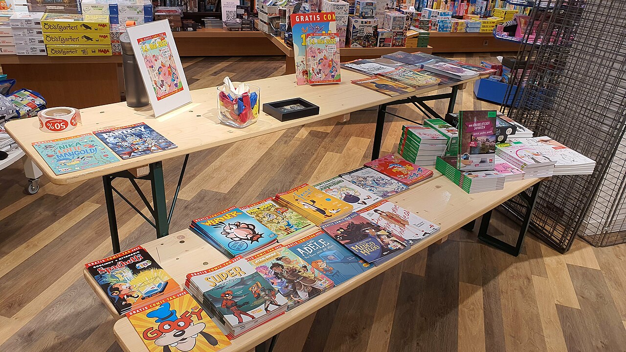 Display of children’s books and educational materials on wooden tables, colorful covers featuring illustrations and characters, arranged neatly in a bookstore or learning center, wooden floor beneath, shelves with more items in the background