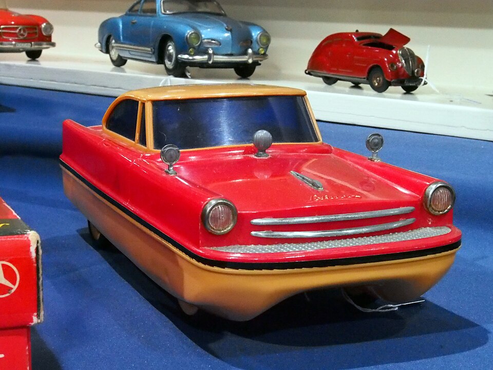 Red and orange toy car with a vintage design on display, blue platform beneath, other colorful toy cars including a blue and red one in the background, all showcased indoors.